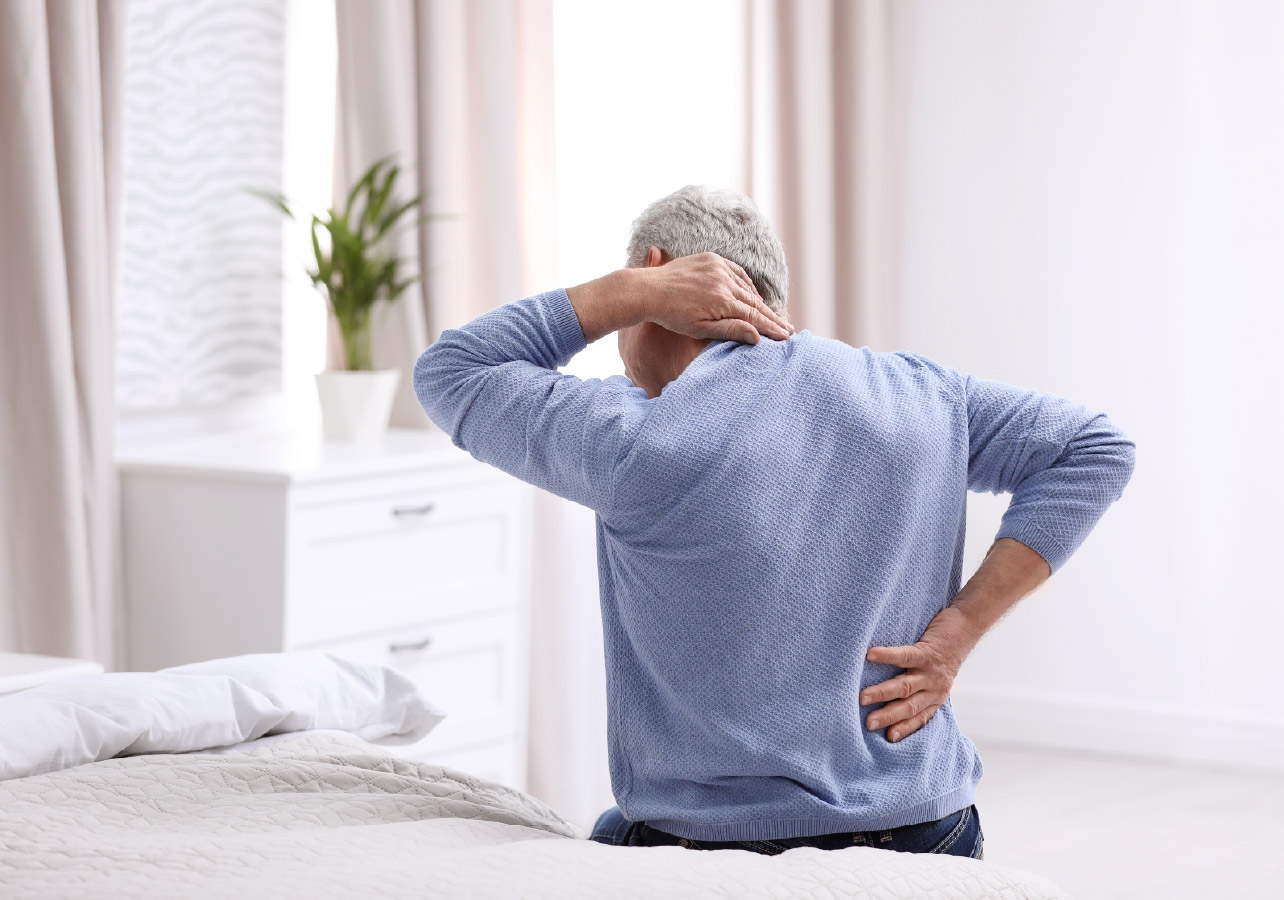 Elderly man sitting on his bed holding his neck and lower back