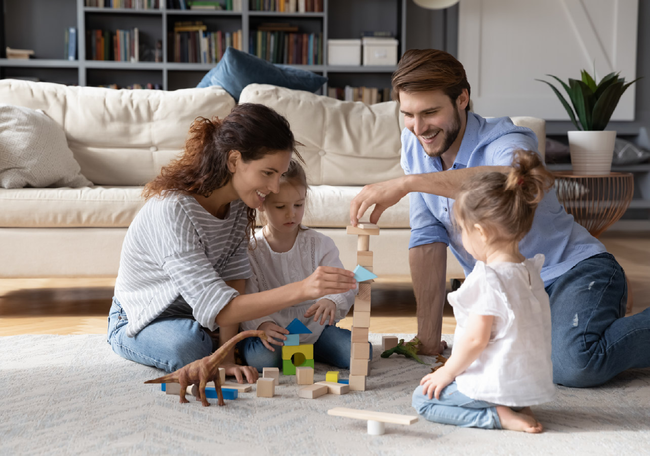 Family of two young adults and two female young children playing blocks on the floor in their living room