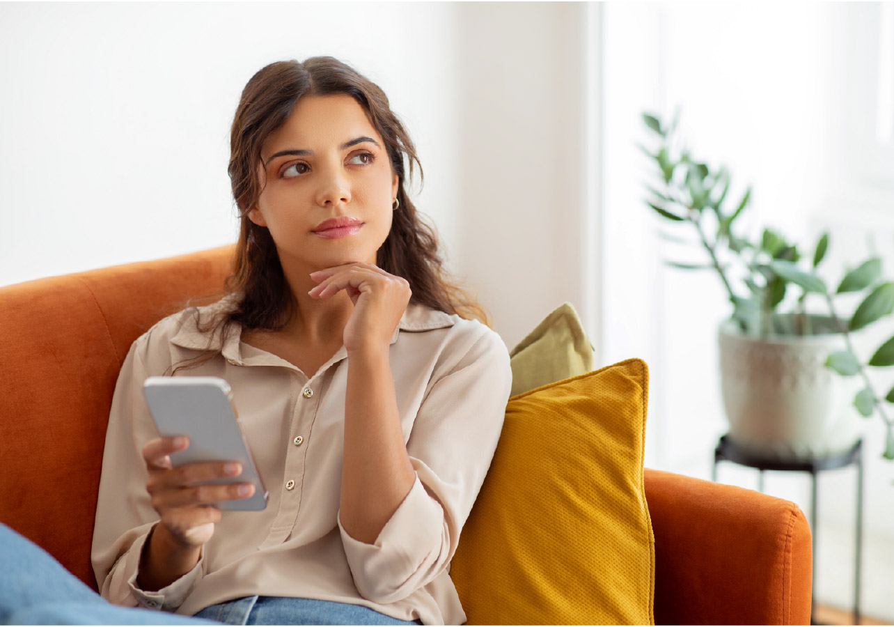 A young woman sitting on a couch, holding her phone and looking into empty space.