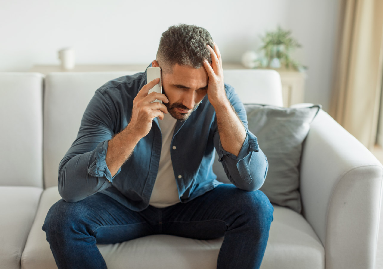 BlogSocialPosts_Nov2025_CC_Ver1_HiddenRisks_BlogHeader Man sitting on a couch holding his phone up to one ear and holding his head with the other hand