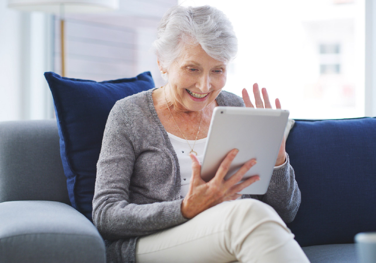 Senior woman sitting on her couch and waving at an IPad she is holding