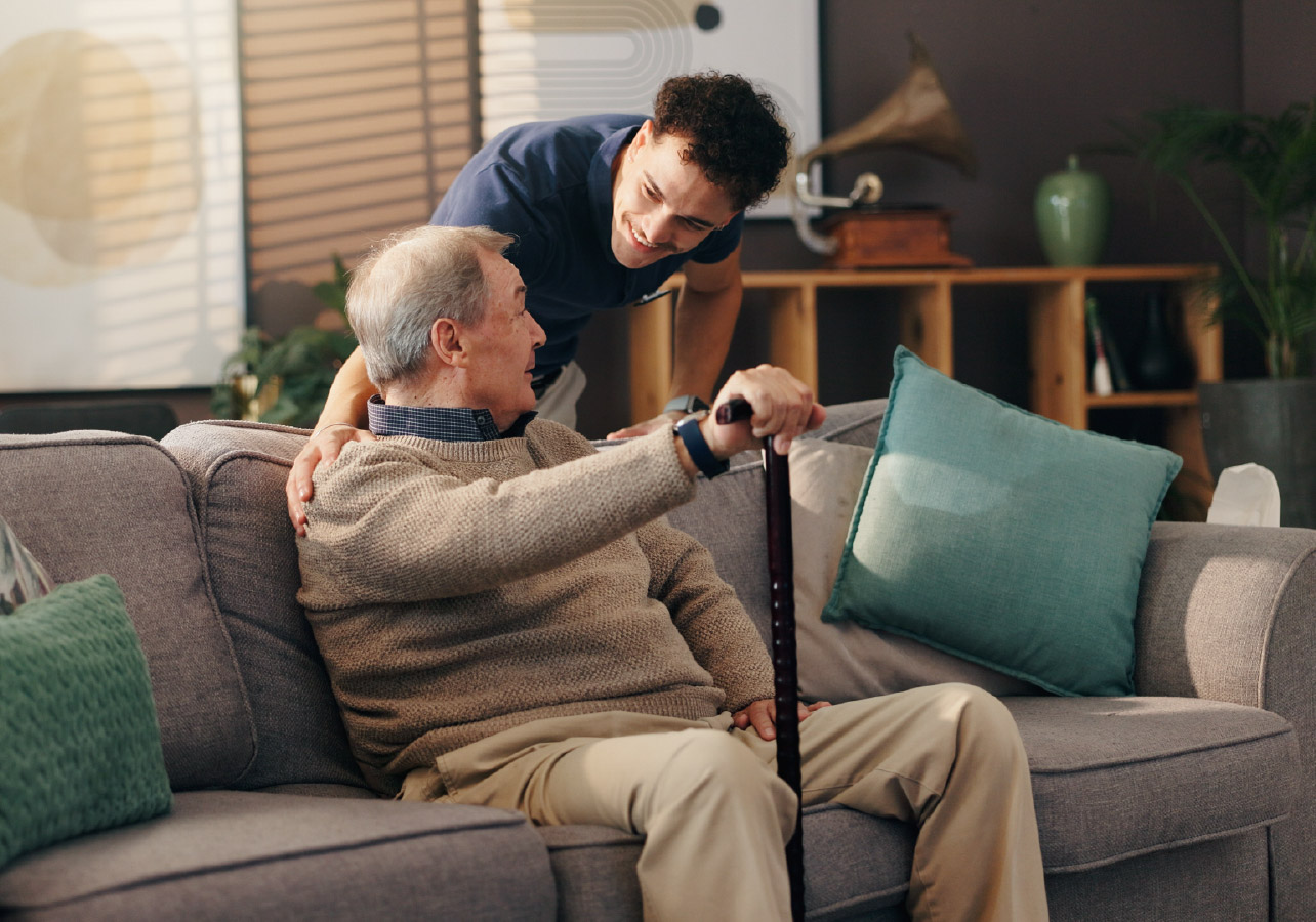 Elderly man sitting on a couch holding a cane with a younger man leaning over the back of the couch talking to him